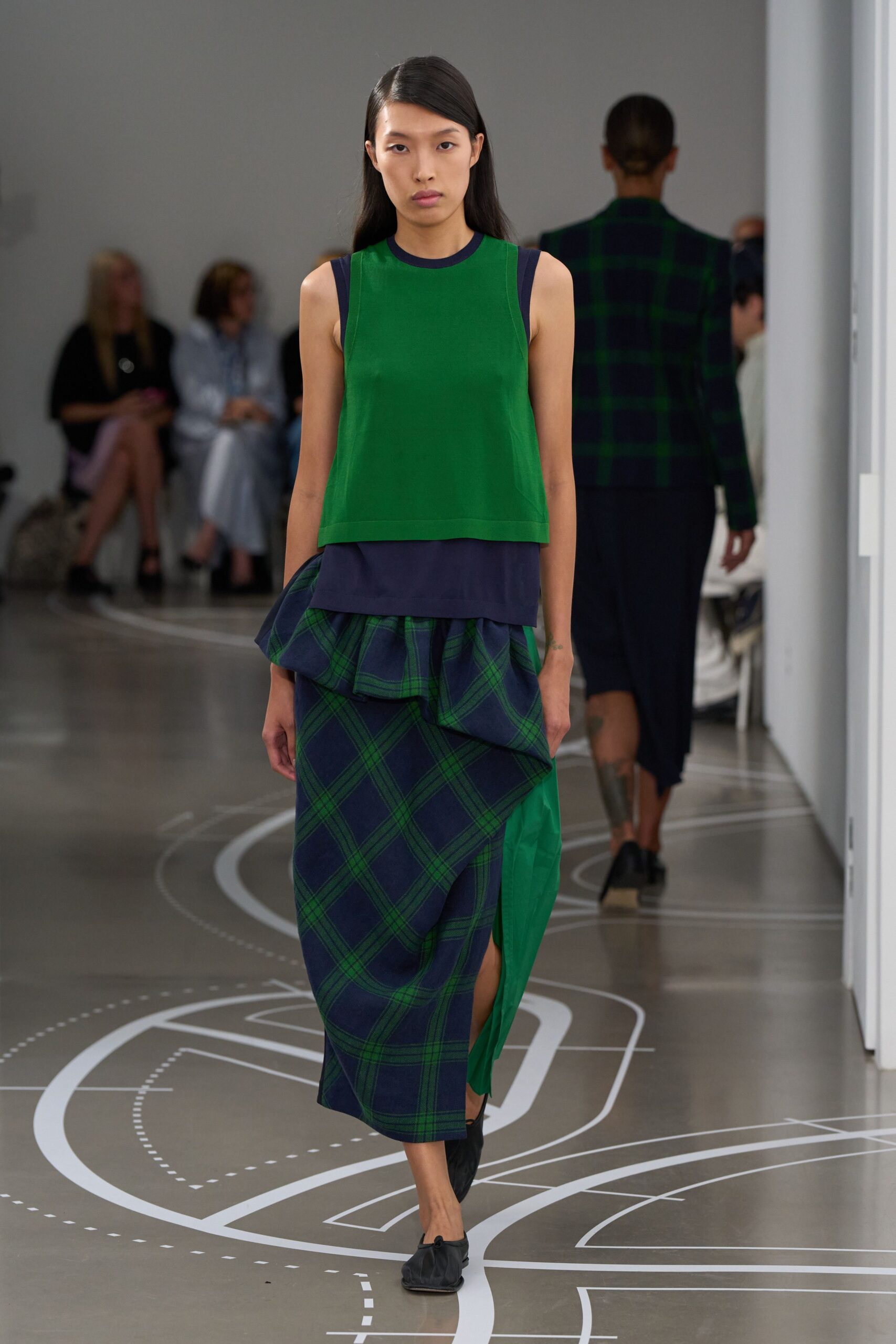 Grass-green knit shell with a narrow navy bound neckline. Navy jersey hem extending below the top. Navy plaid wrap skirt, one side breaking into a bright green panel. Black gathered flats, soft as house slippers. Walking museum corridors after the map folded itself into creases.